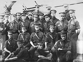a group of male officers wearing dark uniforms, Sam Browne belts and peaked caps sitting and standing on the deck of a ship, a ship gun barrel is visible in the top left of the frame