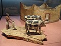 Man holding a yoked plow behind two cows. The cows appear to be yoked together and assisting the man in plowing. In the background a rectangular model of a sandstone building possibly a home is present.