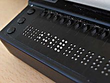 Die Nahaufnahme des linken Endes einer Braillezeile zeigt das schwarze Kunststoffgehäuse, links drei Funktionstasten, daneben einige 8-Punkt-Braille-Elemente. Diese zeigen mit ihren teilweise deutlich sichtbar angehobenen weißen Stößeln folgende Zeichen: zwei Leerzeichen, dann die Zeichen P56, P136, P123 und P12345678 und weitere Leerzeichen.