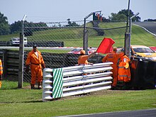 a group of people in fluorescent boilersuits stand behind a barrier, with one of them waving a red flag over the top of the barrier
