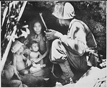 Women with children and dog sitting in cave on left facing a marine squatting on the right who is looking at her.