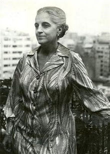 B&W 3/4 length portrait or an older woman wearing a patterned dress, hair in an up-do, standing on an outdoor balcony