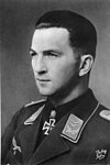 The head of a young man, shown in semi-profile. He wears a military uniform with an eagle above his right breast pocket, an iron cross is displayed at the front of his shirt collar. His hair is dark, short and combed to the back, his nose is long and straight, and his facial expression is determined; looking to the right of the camera.