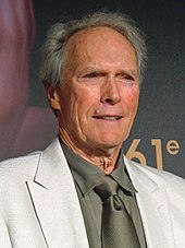 An older man is at the center of the image smiling and looking off to the right of the image. He is wearing a white jacket and a tan shirt and tie. The number 61 can be seen behind him on a background wall.