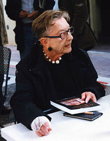 an older lady sitting at a table with a book in her hand