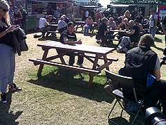 TT rider John McGuinness awaiting a press interview at a picnic bench in the public catering area at the rear of the TT Grandstand during 2013 TT races, with Mrs McGuinness standing to left