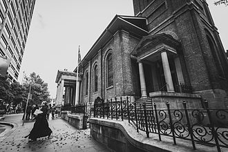 Black and white photo of the rector in black clerical garb striding across the footpath towards Phillip Street