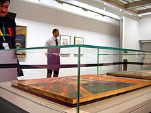 Farbfotografie von einer Seitenaufnahme einer Ausstellungsvitrine, in der das Werk „Der Schrei“ aus dem Jahre 1910 liegend ausgestellt ist. Im Hintergrund stehen ein Besucher und ein Museumswärter. An der hinteren Wand sind weitere Gemälde zu sehen.