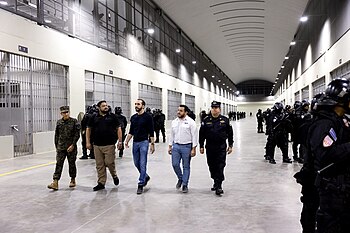 Minister of Defense René Merino Monroy, Director of Penal Centers Osiris Luna Meza, President Nayib Bukele, Minister of Public Works Romeo Herrera, and Director of the National Civil Police Mauricio Arriaza Chicas touring a cell block at CECOT while walking past prison guards wearing riot gear