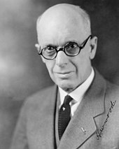 A photographic portrait of a bald white man with thick-rimmed glasses