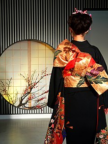 The back view of a young woman, her dark hair tied up, wearing a black furisode and a gold obi with a design of leaves in purple, green and red.