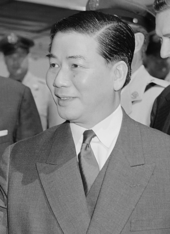A portrait of a middle-aged man, looking to the left in a half-portrait/profile. He has chubby cheeks, parts his hair to the side, and wears a suit and tie.