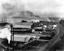 A black-and-white photograph of several factories on a waterfront, with a railroad running past them.