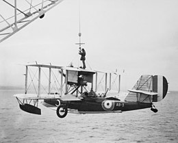 Single-engined military biplane with floats under wings being hoisted out of the ocean by a crane. A crewmen is riding on its top wing, holding the crane's cable.