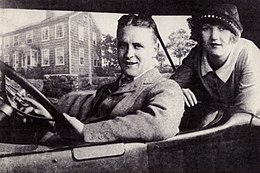 A black and white photograph of F. Scott Fitzgerald and Zelda Fitzgerald seated in a convertible vehicle. A smiling Scott is behind the steering wheel and wears a tweed suit. Zelda is in the rear seat behind Scott wearing a Cloche hat and a Peter Pan shirt collar. Her right arm is resting on the back of the driver's seat.