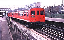A three-quarter photograph of a red train O stock with sliding doors and flared sides, leaving Barking station bound for Ealing in 1980