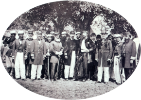 A photograph showing a large group men, most of whom wear uniforms, standing beneath shade trees