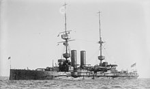 A large, dark gray warship bristling with guns sits at anchor