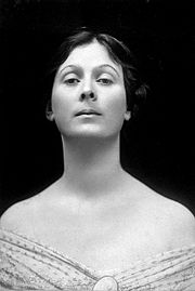 A black and white studio photograph of American dancer Isadora Duncan staring at a downward angle with an imperious air. Her shoulders are bare, and her dress has a decolletage neckline.