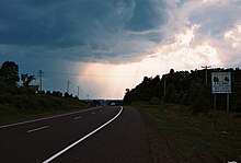a highway curving over the crest of a hill flanked by telephone poles on either side. There is a white road sign on the right side of the road and storm clouds in the sky.