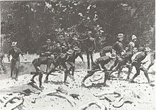 Black and white print of an oil painting by Frederic Remington shows slouch-hatted soldiers advancing in a crouch while crossing a stream