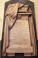 Photograph of a marble memorial on a wall with carved fabric draped over it and text on a panel