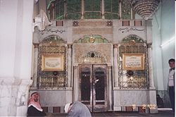 The interior of a religious building showing two men seated in front of a gated marble enclosure covered in green-tinted glass