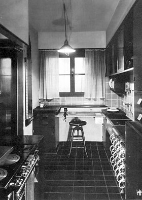 Black and white photograph of a narrow kitchen with a window over the sink, plus a stool, built-in storage, and adjustable lighting