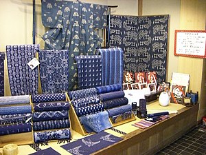 A display table showing a number of different indigo-dyed kasuri fabrics, some stacked on wooden stands in roll-form, others displayed flat in front of the rolls, and some hanging on small stands.