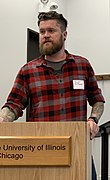 American man wearing a slim-fitting flannel shirt, a beard, and an undercut, 2019. Sleeve tattoos can be seen.