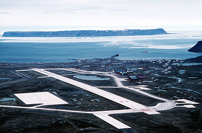 An aerial view of Pituffik Space Base in Greenland, taken in the 1980s when it was named Thule Air Base, showing a large runway and surrounding track, and a coastal town of one-storey buildings, with two ships backgrounded by sea ice.