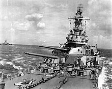 A large ship tilted to the right, with gun barrels pointed to the left. Crewmen can be seen on the battleship's deck. To the left of the image another large warship can be seen.