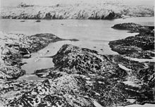 Black and white aerial photo of a body of water surrounded by steep snow-topped mountains. Several ships are anchored in a bay, with the battleship Tirpitz's location being marked with an arrow.