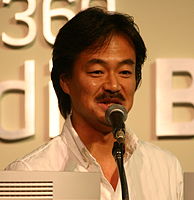 A 43-year-old Japanese man with neck-length black haired, speaking into a microphone and facing slightly to the camera's right.