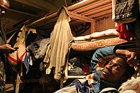 A refugee man is lying on a shelf. There are multiple shelves in the background on which there are blankets. The shelf serves as the bed for the man.