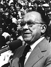 portrait from the chest upwards of man giving speech, a large crowd slightly out of focus is behind him