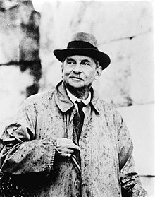 Portrait of Evans: an elderly man in a raincoat and hat, in front of an ancient stone wall.