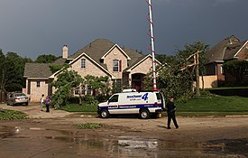 A news van with a microwave antenna fully extended in front of a section of tornado-damaged houses.