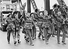 Black and white photo of a group of men wearing military uniform walking on a road in front of a bus