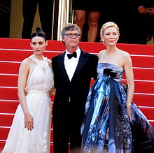A brunette woman in a white dress, a man in a tuxedo, and a blonde woman in a dress that is mostly blue with other colored patterns stand in front of red stairs