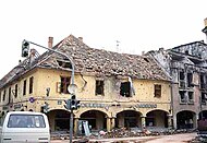 Two shattered arcaded buildings, one old and painted yellow, the other more modern and made of brick and concrete, with destroyed roofs and many bullet holes. In front is a damaged traffic signal and a car.