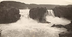 Black and white photograph of two parallel waterfalls, dropping over a dark cliff face into a turbulent pool.
