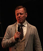 Abel Korzeniowski, standing and speaking into a hand-held microphone at a film-music festival