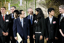 President George W. Bush and Japanese Prime Minister are on the right of a high school student. The high school student speaks. In the background are more high school students and important political figures.