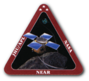 An artwork of a spacecraft hovering above an asteroid, enclosed in an equilateral triangle with a thick, red border. The words "JHU/APL", "NASA", and "NEAR" are printed in bold white font, on the left, right, and bottom sides of the triangle's borders.
