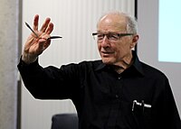 Elderly man in black shirt and glasses holding up a bent spoon by the neck
