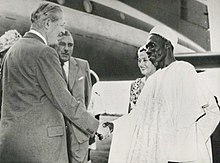 A white man in a business suit shakes hands with a black man in white robes.