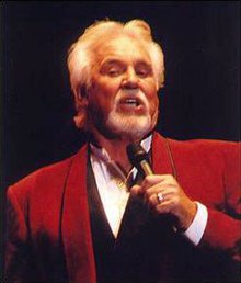 A man with a gray mustache, beard, and hair, wearing a red jacket and singing into a microphone