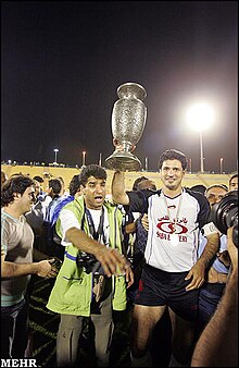 Photograph of one player holding the trophy above his head and smiling at the camera while standing among a crowd of other players and officials