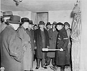 Buchenwald survivor explains to a visiting delegation some brutal methods of keeping order in the camp; he wears a Lagerdienst ("camp service", prisoner-functionary helper of the guards) armband.